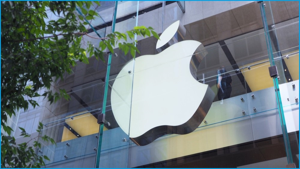 A close up of a large Apple logo hovering behind glass at the Apple Store in Sydney's CBD, with leaves from a tree in the foreground.